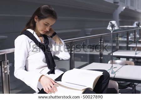 Pretty student sitting on desk in modern university library studying a book.