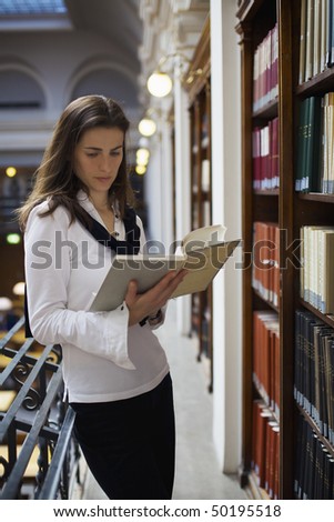 Young attractive student standing at bookshelf in old university library reading a book.