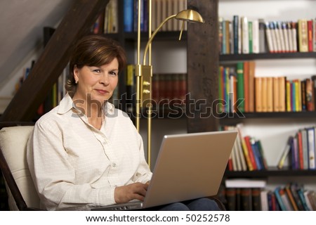 Modern senior woman sitting in front of bookshelf and working on laptop.