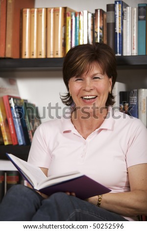 Laughing senior woman sitting on floor in front of bookshelf at home and reading a book.