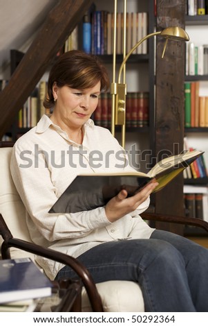 Pretty senior woman sitting in chair at home in front of bookshelves reading a book.