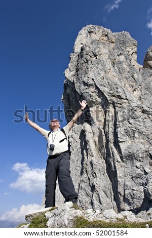 Active senior hiker standing in front of huge rock formation near summit lifting his arms out of joy to be in the Alps, Tyrol, Austria.
