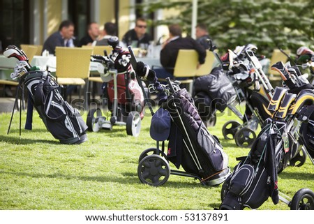 Several golf bags parking at club house with group of golfers sitting in background.