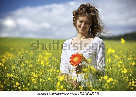 Happy woman in white dress holding red flowers in yellow rapeseed field.