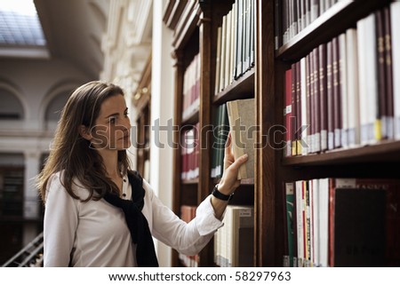 Pretty female student standing at bookshelf in old university library searching for a book.