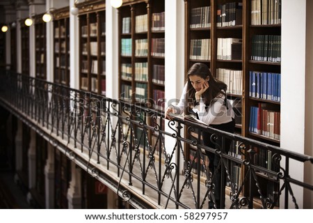 Female student leaning at handrail in old library reading a book; bookshelf with old books collection in background.