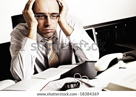 Worried businessman sitting at office desk full with books and papers being overloaded with work.