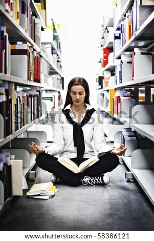 Female student sitting in Yoga pose in between bookshelves in modern university library and meditating over a book.
