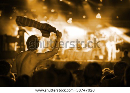Fan on shoulder of another person at live pop concert cheering at band and swinging his T-shirt.