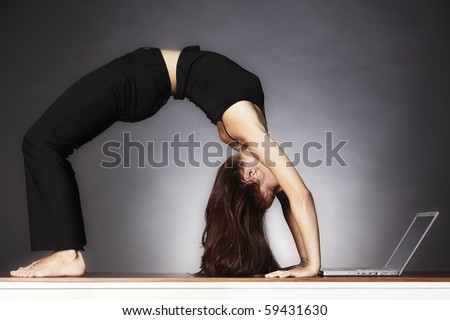 Young pretty woman in yoga wheel posture looking at laptop, backlit grey background.