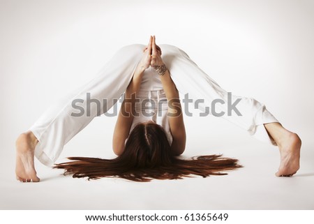 Young lady practicing yoga in variation of plough posture (halasana) in white clothes on white background, high-key image.