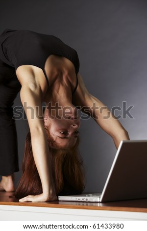 Woman in yoga wheel posture looking at laptop, backlit grey background.