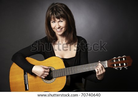 Smiling girl in black sitting and playing on guitar looking straight, isolated on black background.