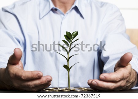 Businessman in blue shirt protecting with hands green plant growing out of coins pile symbolizing safe growth of financial wealth.