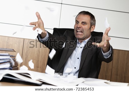 Overloaded senior businessman being upset about work, tearing papers and screaming, sitting at office desk in front of many books and files.