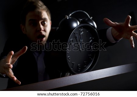 Young desperate business person in dark suit at office desk reaching alarm clock showing five minutes to twelve o'clock and trying to rescue his business, low-key image isolated on black background.