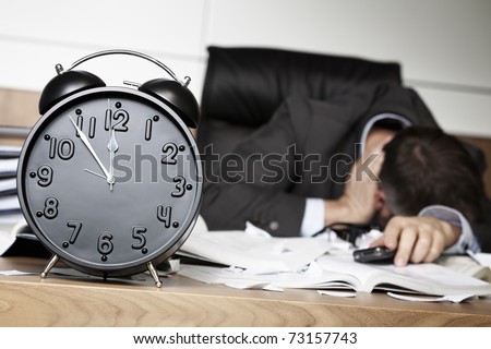 Senior business person being shocked after receiving bad news on cell phone, sitting at office desk behind alarm clock showing five minutes to twelve.