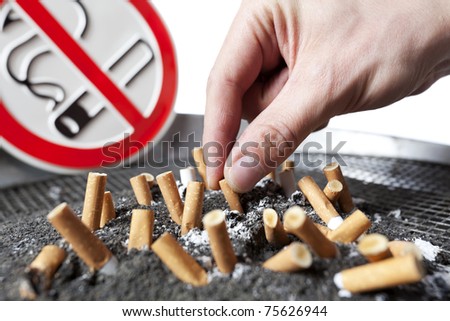Close up of human hand with cigarette butts stuck in ash with no smoking sign in background.