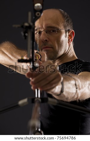 Close up of handsome bowman in black on black background aiming with bow and arrow, front view with focus on eyes.
