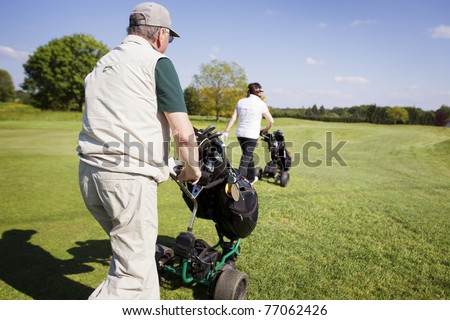 Active senior golf couple pushing golf trolley with bag on fairway on beautiful golf course with blue sky in background.