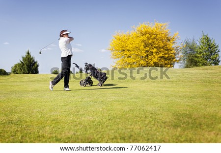 Active senior female golf player swinging golf club to shoot ball on fairway on beautiful golf course with blue sky in background.