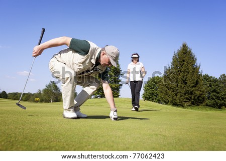 Senior active male golf player picking up golf ball from hole on green with female golf player walking in background with fantastic blue sky.