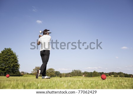 Active senior female golf player swinging golf club to tee off ball on beautiful golf course with blue sky in background.