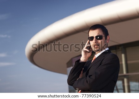 Close up of young cool business person in black suit and sunglasses being busy on cell phone with office building and blue sky in background.