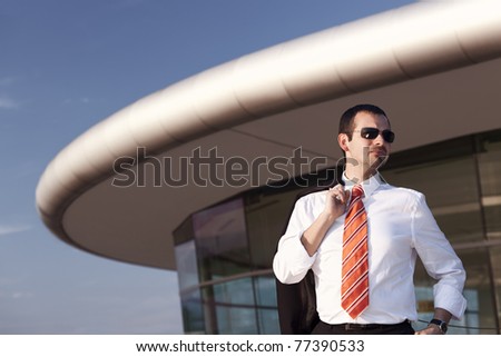 Serious young business person in white shirt, orange tie and sunglasses standing in front of office building with blue sky in background.