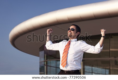 Successful business person in white shirt, orange tie and sunglasses raising his hands in front of office building and blue sky.
