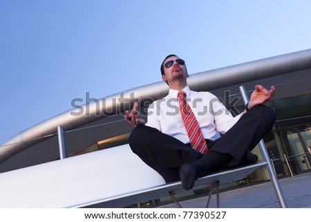 Calm balanced business person sitting in Yoga pose on bench meditating with office building in background.