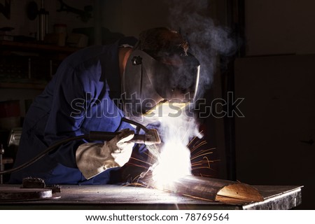 Industrial electrode welder with face shield and blue overall welding a piece of metal in workshop.