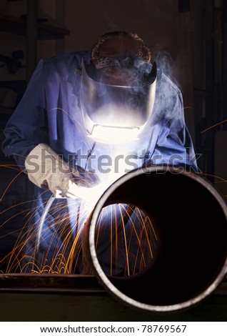 Industrial electrode welder with face shield and blue overall welding a steel pipe in workshop.