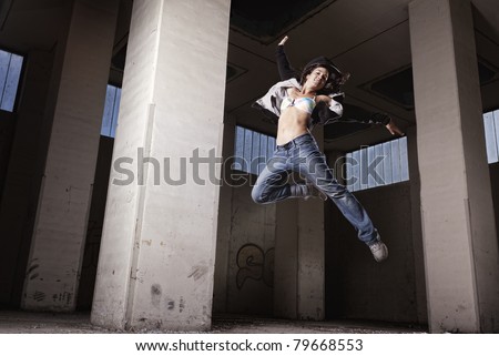 Smiling sexy modern dance girl jumping high with one leg up in old grungy factory hall.