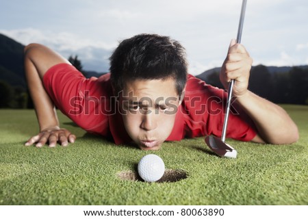 Desperate young male golf player in red shirt and putter lying on golf green and blowing golf ball into cup, focus on ball.
