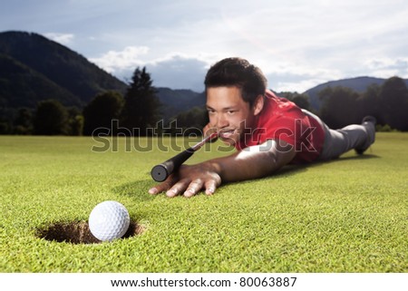 Happy young male golf player lying on green and using golf club as billiard cue (queue) to hole ball into cup.