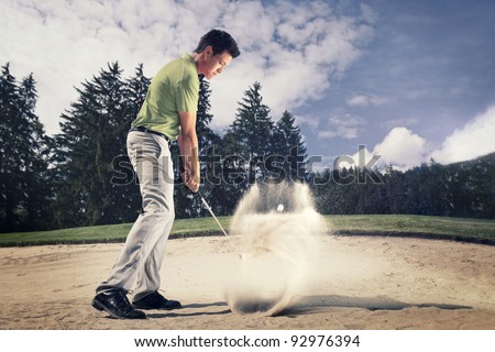 Male golf player in green shirt and grey pants hitting golf ball out of a sand trap with sand wedge and sand caught in motion.