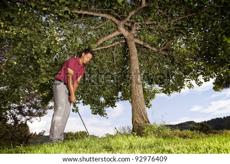 Male golf player underneath a tree in red shirt and grey pants focusing on golf ball to hit with iron.