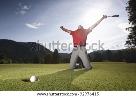 Male golf player on knees and arms raised with putter in hand in winner pose on golf green being overjoyed as golf ball drops into cup.