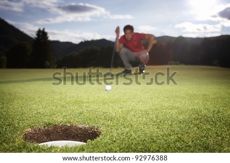 Male golf player in red shirt squatting to analyze the green for putting the golf ball into the hole.