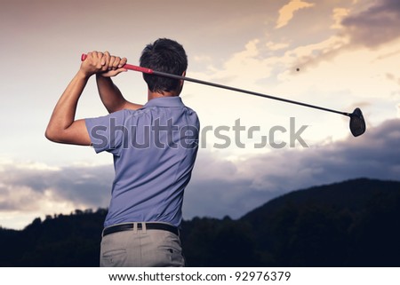 Close up of professional golf player in blue shirt teeing-off ball at sunset, view from behind.