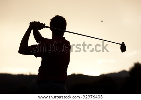 Silhouette of golf player teeing-off ball at sunset, view from behind.