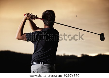 Close up of male golf player in black shirt teeing-off ball in twilight, view from behind.