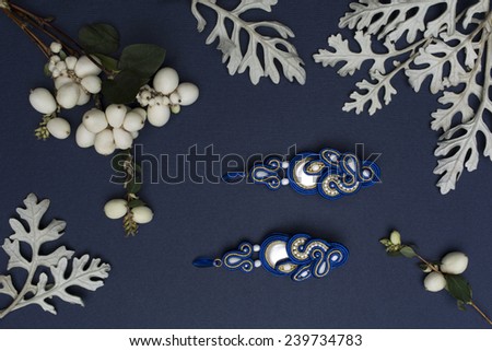 Soutache bijouterie blue earrings with white and blue crystals on dark blue background with white leaves and berries