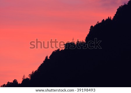 A house perched at the ridge of a tree covered mountain slope against dramatic dark read evening sky, rendered as silhouette with lit windows.