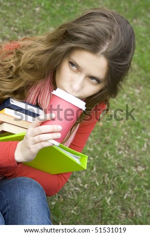 Young beautiful student at a dinner in the park sits on the green grass drinks coffee. Keep lots of books and folders