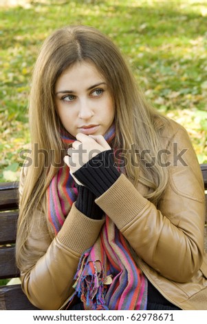 Vertical portrait of a beautiful young woman sitting on a bench in the park around a lot of yellow, red green leaves. Breath warms your hands