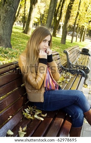 Vertical portrait of a beautiful young woman sitting on a bench in the park around a lot of yellow, red green leaves. Shivering, wrapped in a jacket