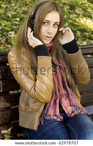 Young Caucasian woman with headphones in autumn park sitting on a wooden bench, listening to music and singing. Autumn around a lot of colorful foliage