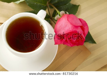 White cup of tea next to sugar and is a beautiful red rose lying on sheets of old paper sheet music